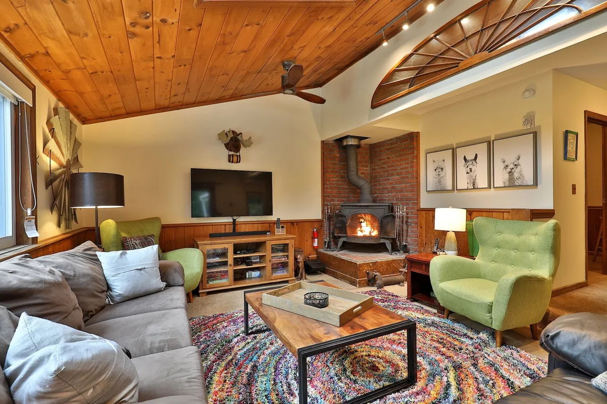 This warm and inviting living room features a stunning wood-beamed ceiling, a crackling fireplace, plush green armchairs, and a colorful rug—all arranged for guests to gather, relax, and unwind after a day on the slopes.