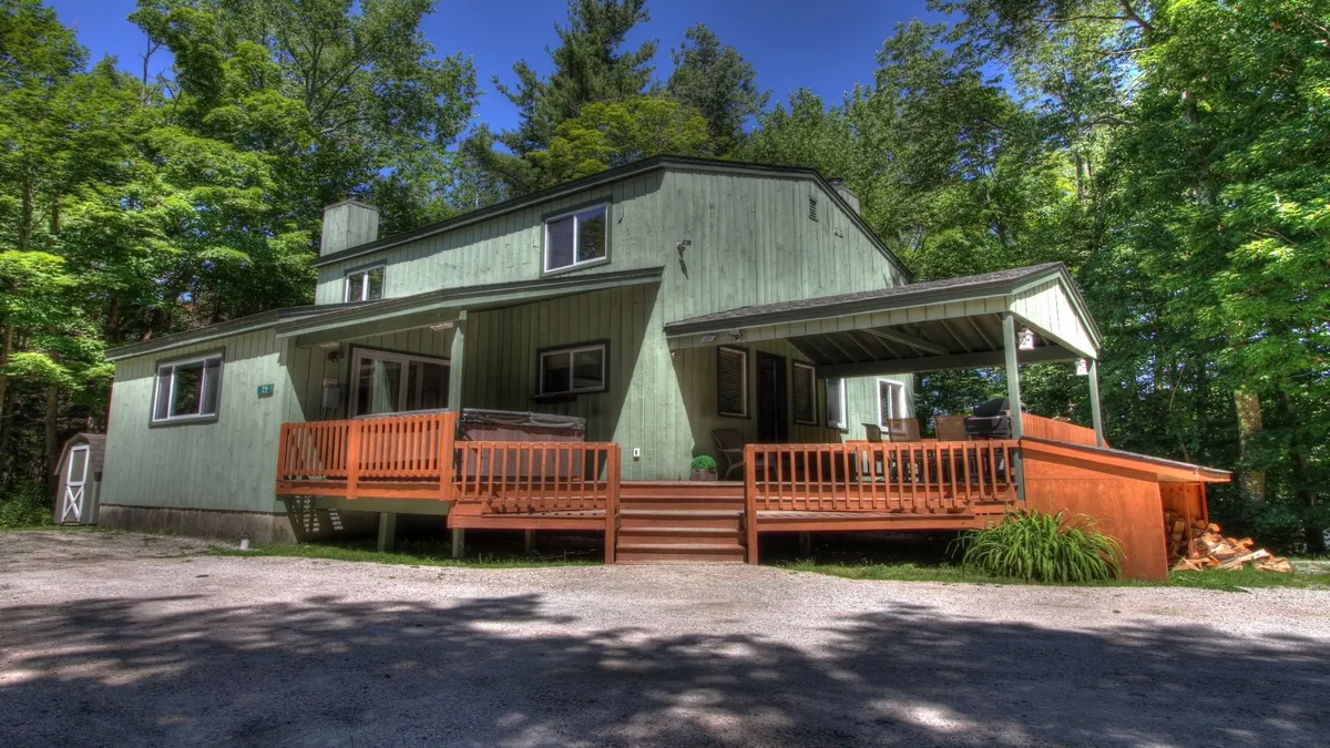 This image displays the green wooden exterior of the house, a spacious deck surrounded by mature trees, and a stack of firewood. The deck is set up for outdoor dining and relaxation, inviting guests to enjoy the natural surroundings and communal gatherings.