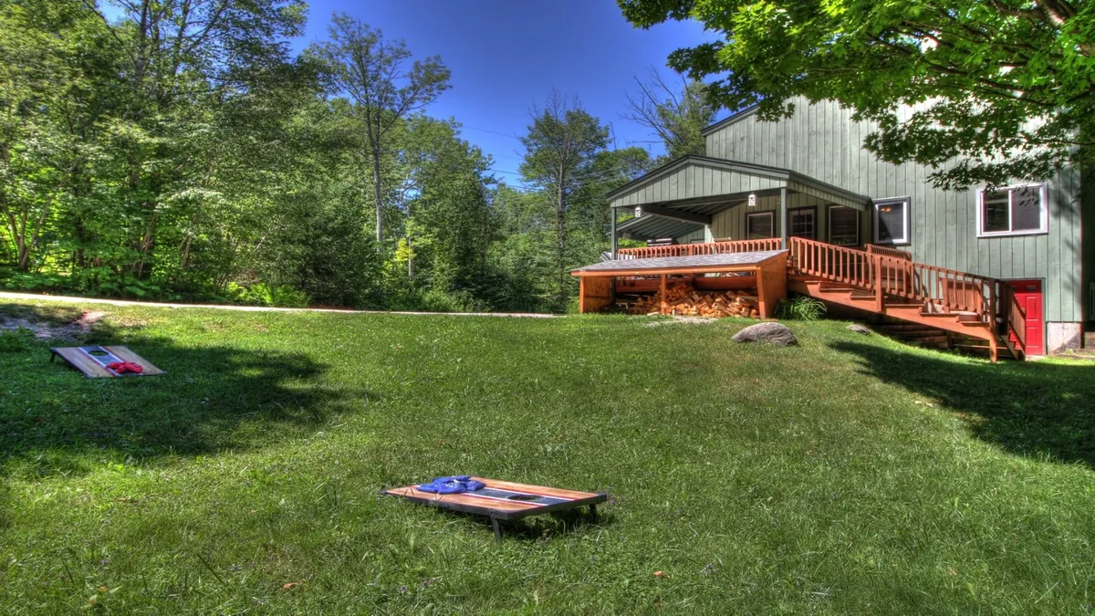 This exterior image showcases the house, its deck, a grassy area, and cornhole boards. The setting hints at space for group gatherings and outdoor play, with a welcoming, playful mood.