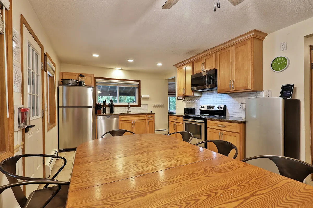 A cozy interior scene featuring a large dining table, kitchen appliances, and windows with scenic views. The space is designed for socializing, with ample seating for group meals and family celebrations.