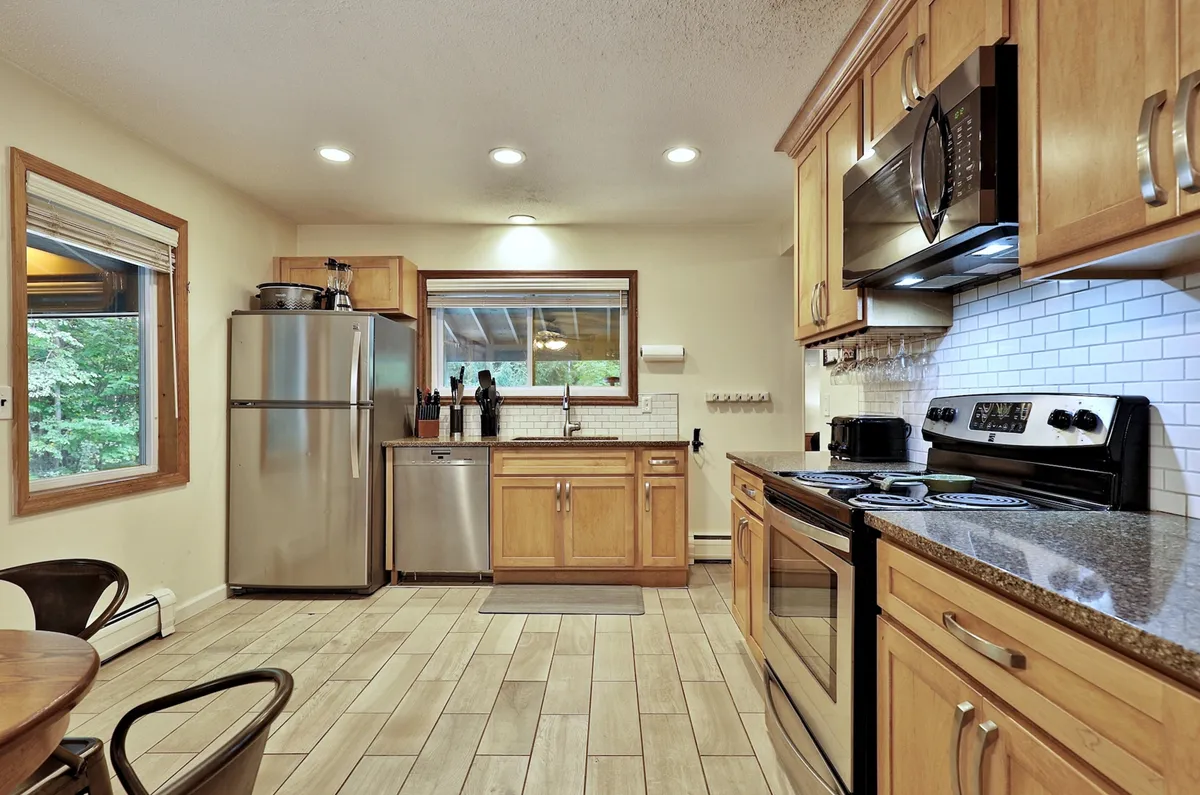 A modern kitchen scene with stainless steel appliances, wooden cabinetry, a large window that floods the space with natural light, and granite countertops. The setup is perfect for cooking, entertaining, and hosting group feasts with all necessary amenities visible.