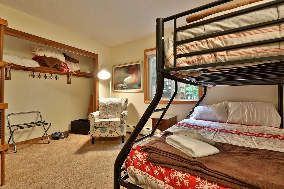 A welcoming bedroom with a sturdy twin-over-full bunk bed, an inviting armchair for reading, cheerful wall art, and a closet with open shelves. The room is well-lit and arranged for both sleep and relaxation, embodying the cozy, communal feel ideal for families or groups traveling together.