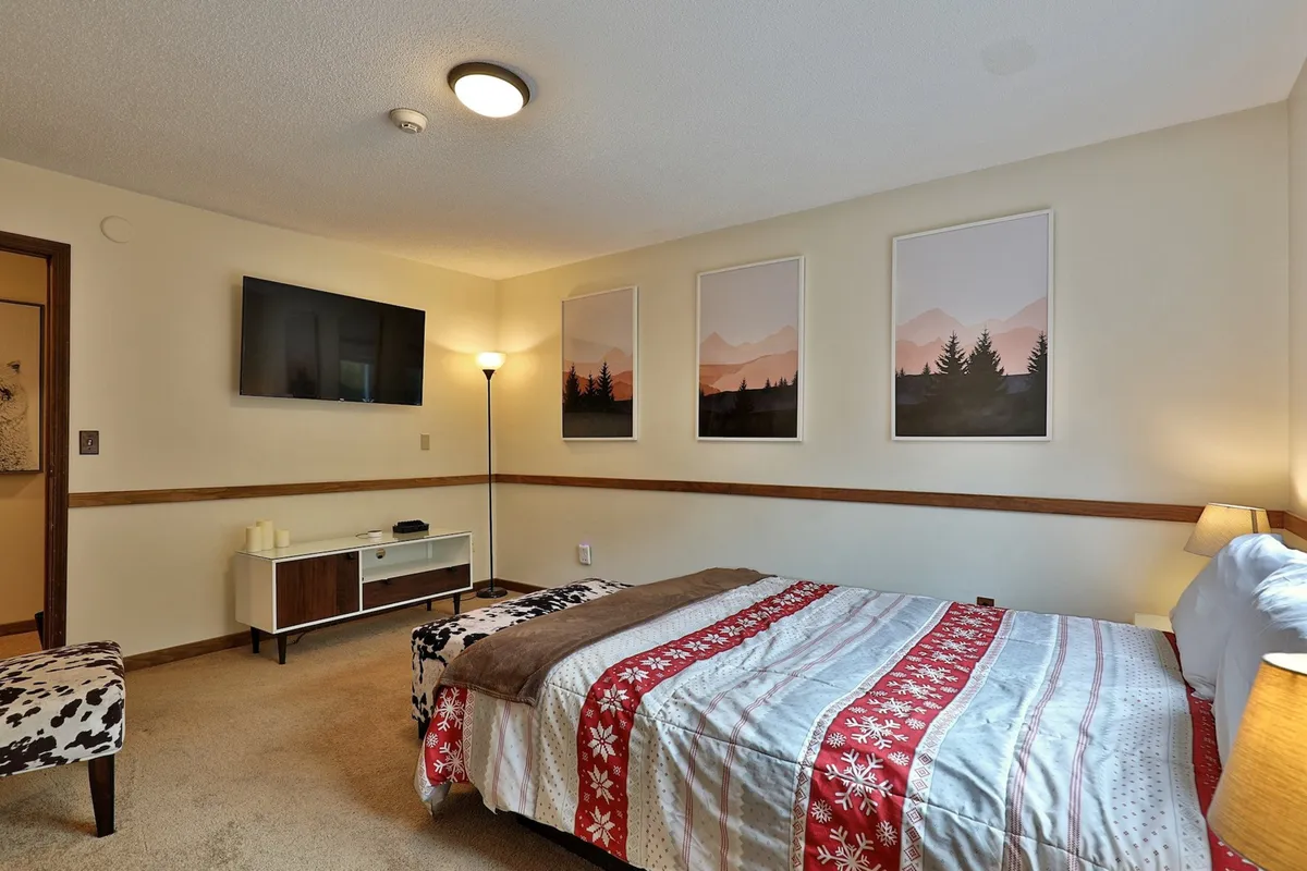 A serene bedroom featuring a large bed with a patterned blanket, a wall-mounted smart TV, landscape artwork, a floor lamp, and wooden furnishings. The ambiance is tranquil, ideal for unwinding after an active day, with amenities that suggest both convenience and relaxation.