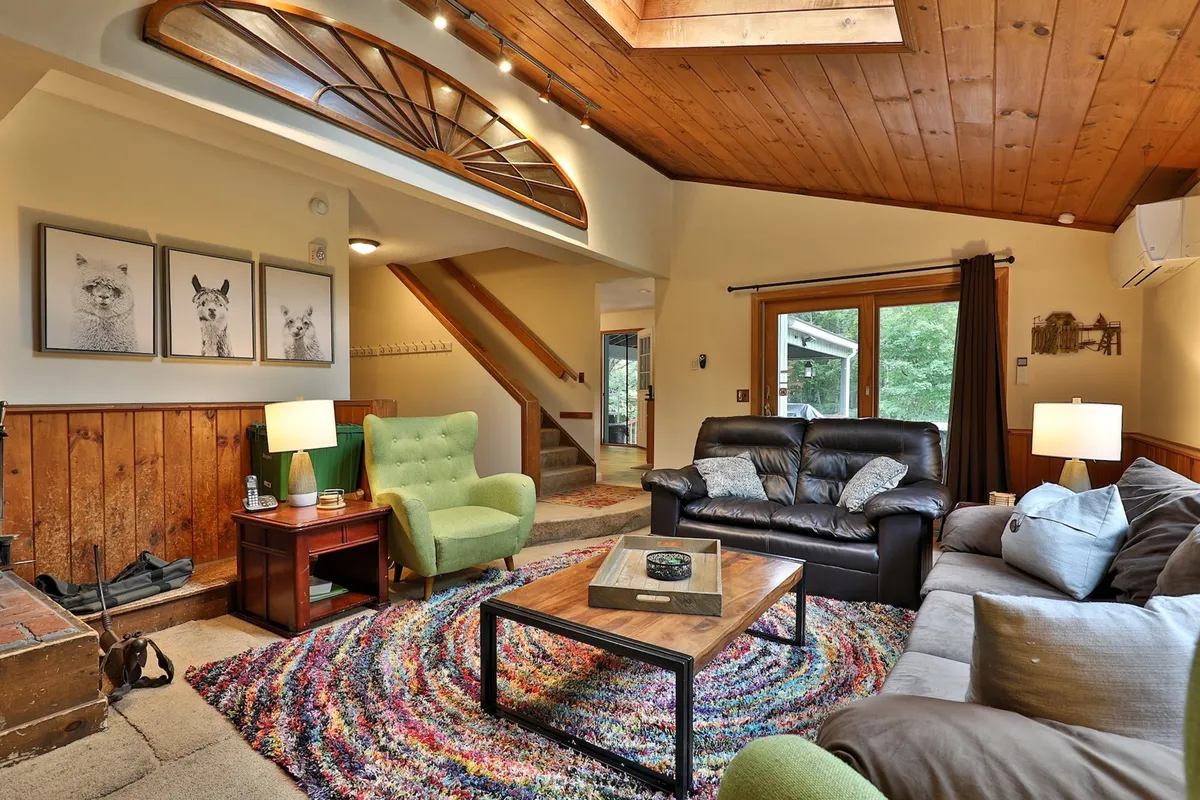 A vibrant, inviting living area featuring a colorful rug, green armchair, framed animal art, and prominent wood accents. The space is well-lit with natural sunlight coming through the windows, creating a comfortable atmosphere ideal for both relaxation and casual socializing.