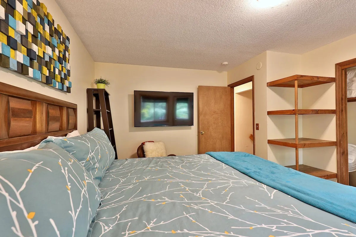 A restful bedroom scene with a bed adorned in patterned bedding, colorful wall art, a TV mounted on the wall for entertainment, and an open shelving unit for storage. The space feels calm and organized, suitable for winding down after a productive day.