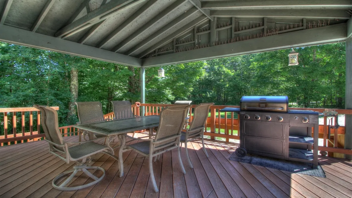 The exterior deck features a BBQ grill, a dining table, wooden railings, and lush surrounding trees. The setup is ideal for outdoor dining, relaxing, and socializing, with a clear view of the natural surroundings and communal layout.