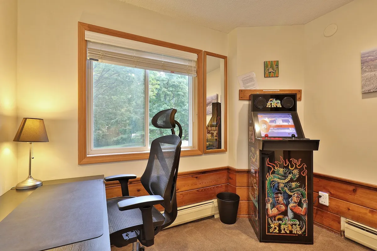 A dedicated entertainment area with a gaming cabinet, desk, window for natural light, and a cozy lamp. The setup is ideal for both group gaming sessions and quiet moments, with a nostalgic atmosphere that appeals to all ages.