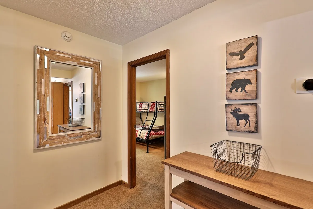 This image captures a cozy entryway with a wooden mirror, artwork on the wall, wooden console table, and an open doorway leading into a bedroom. The warm, inviting decor subtly suggests a space welcoming guests from outdoor adventures.