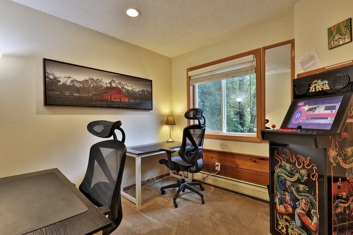 This image showcases a dedicated office space featuring two desks with ergonomic chairs, framed landscape artwork on the wall, a large window filling the room with natural light, and a retro arcade machine in the corner. The arrangement is functional yet inviting, balancing a professional setup with playful touches.